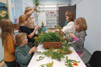 Na stole leży koszyk z kwiatami nawłoci. Dzieci pod okiem instruktorki przeglądają rośliny i wyciągają je z kosza.