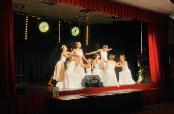 Na scenie teatralnej grupa kobiet w białych sukienkach wykonuje układ taneczny, podnoszą inne tancerki.
