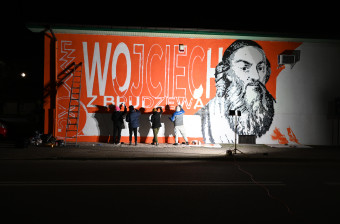W ostrym świetle reflektorów, grupa młodych ludzi wypełnia napisy do muralu z Wojciechem z Brudzewa
