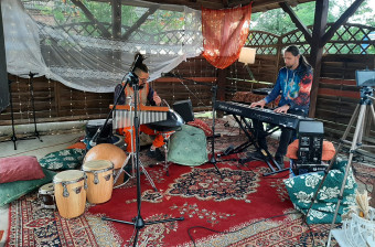 Wiata ozdobiona materiałami i perskim dywanem. W środku siedzi dwóch mężczyzn, jeden gra na syntezatorze a drugi na cymbałach, z boku stoją bębny.